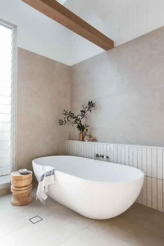 a serene Scandinavian bathroom with large scale tiles, an oval tub, a beam, a stool and some greenery