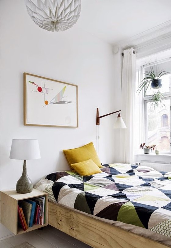 a small Scandinavian bedroom with a bed with bright bedding, a stained nightstand, a sconce and an artwork