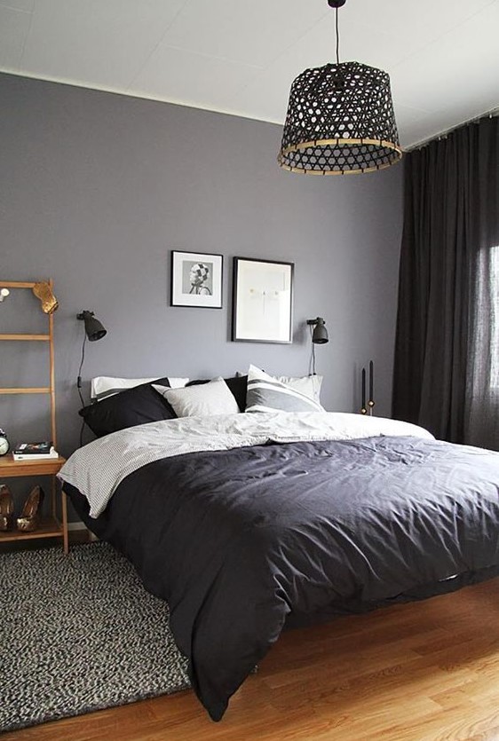 a small moody bedroom with a grey wall, a bed with monochromatic bedding, a woven pendant lamp and black curtains