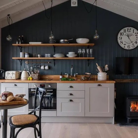 A small pretty kitchen with grey cabinets, a navy accent wall, open shelves, butcherblock countertops and a small hearth.