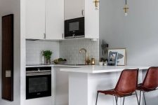 a small white kitchen with white tiles, pendant lamps, brown leather stools and gold touches is chic