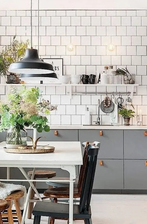 A Scandinavian kitchen features grey cabinets with leather pulls. White square tiles, vintage chairs, and pendant lamps add style. This design combines modern and classic elements.