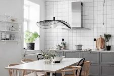 a stylish Scandinavian kitchen with white square tiles, grey shaker cabinets, a hood, a white table and stained chairs and a catchy pendant lamp