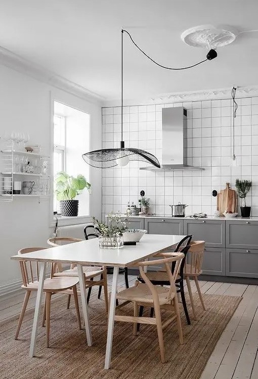 a stylish Scandinavian kitchen with white square tiles, grey shaker cabinets, a hood, a white table and stained chairs and a catchy pendant lamp