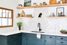 a stylish navy L-shaped kitchen with a white Zellige tile backsplash, open shelves, greenery and black fixtures