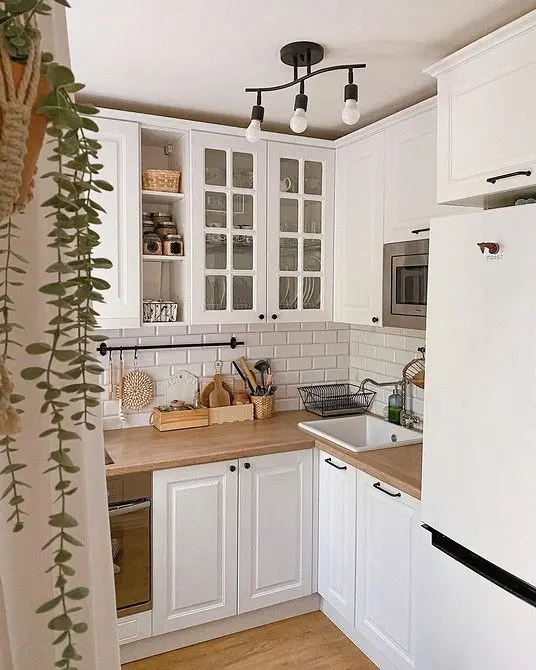 A tiny white L shaped kitchen with a subway tile backsplasj, butcherblock countertops, a modern chandelier and greenery