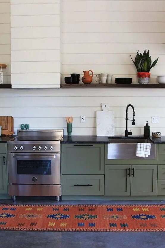 A vintage boho green one wall kitchen with black countertops, a white beadboard backsplash and a hood, a colorful rug.