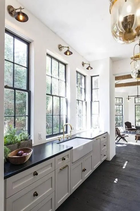A vintage white one wall kitchen with black countertops, large windows, brass sconces and elegant pendant lamps.