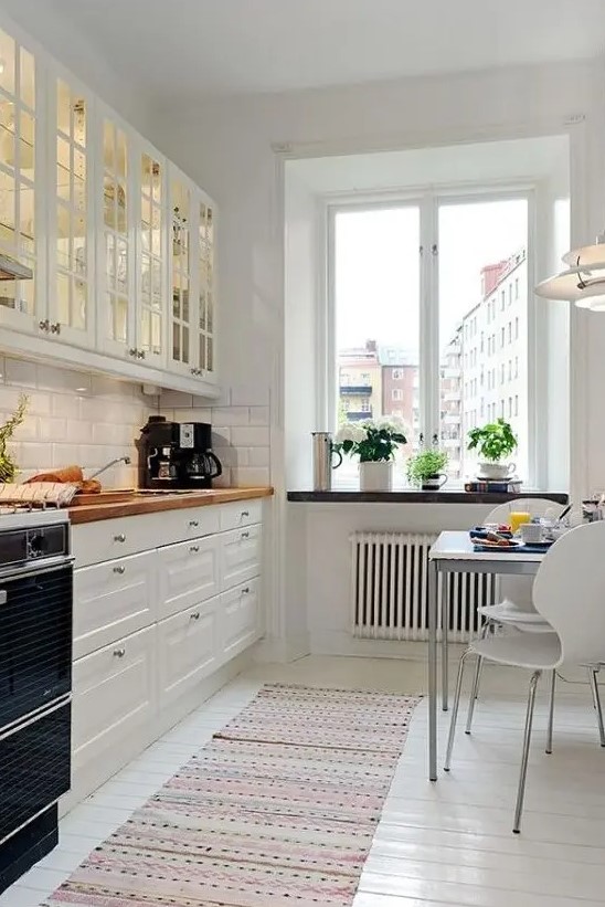 Butcherblock countertops add warmth to this welcoming white Scandinavian kitchen, complemented by glass cabinets and white tiles. A small eating zone offers a cozy spot for meals. Use butcherblock for a practical and inviting kitchen surface.
