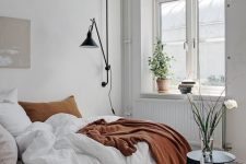 a white Scandinavian bedroom with a low bed, neutral bedding, coffee tables, a black sonce and some plants