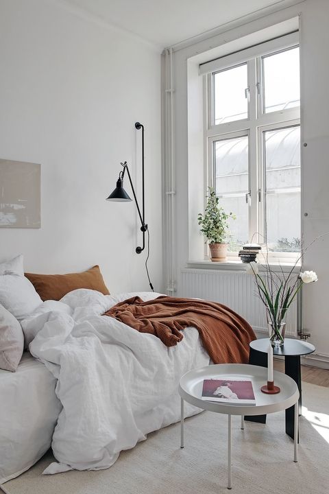 a white Scandinavian bedroom with a low bed, neutral bedding, coffee tables, a black sonce and some plants