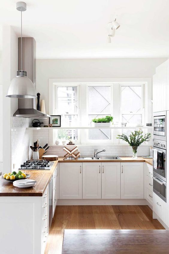 a white farmhouse U-shaped kitchen with butcherblock countertops, built-in appliances, pendant lamps and greenery