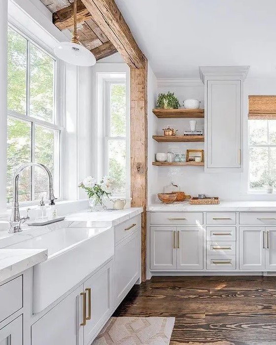 a white farmhouse kitchen with a wooden beam, wooden shelves and woven shades plus brass touches