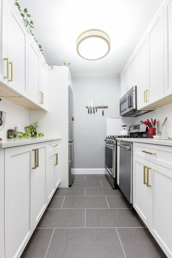 a white galley kitchen with shaker cabinets, white stone countertops, gold handles, a white backsplash and a ceiling lamp