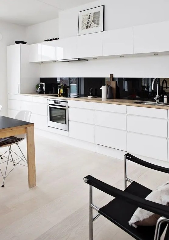 A white minimalist one wall kitchen with blonde butcherblock countertops, a black glass backsplash and an eating zone.