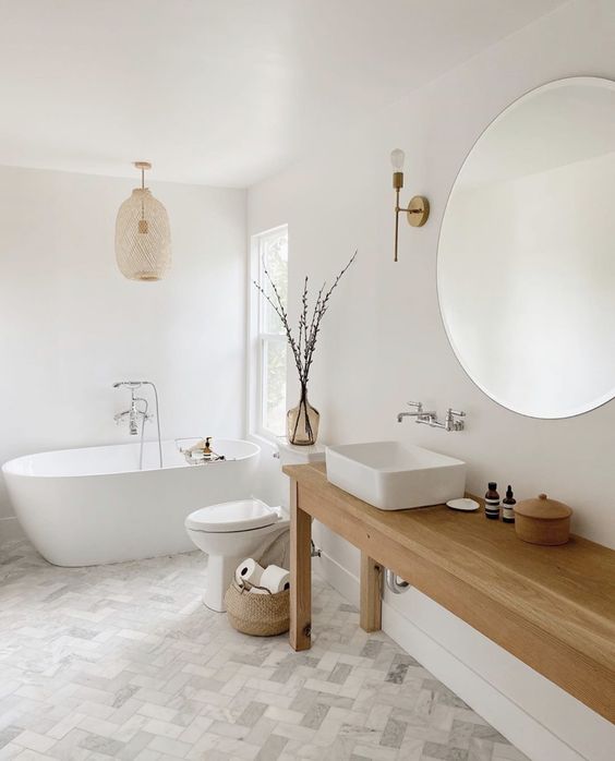 an airy Scandinavian bathroom with herrinbone tile, a wooden table as a vanity, a sink, a round mirror and lamps