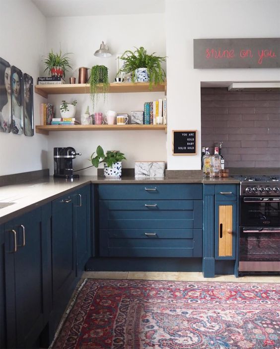 an eclectic kitchen with navy cabinets, open shelves, a bright rug, a mauve brick backsplash, some decor and greenery