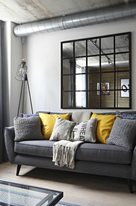 an industrial living room with white walls, exposed pipes, a grey sofa, grey and mustard pillows, a framed mirror and an industrial lamp