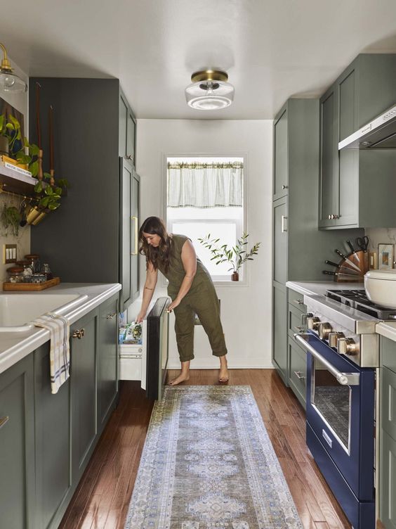 an olive green galley kitchen with a navy cooker, white countertops, some greenery and a small window