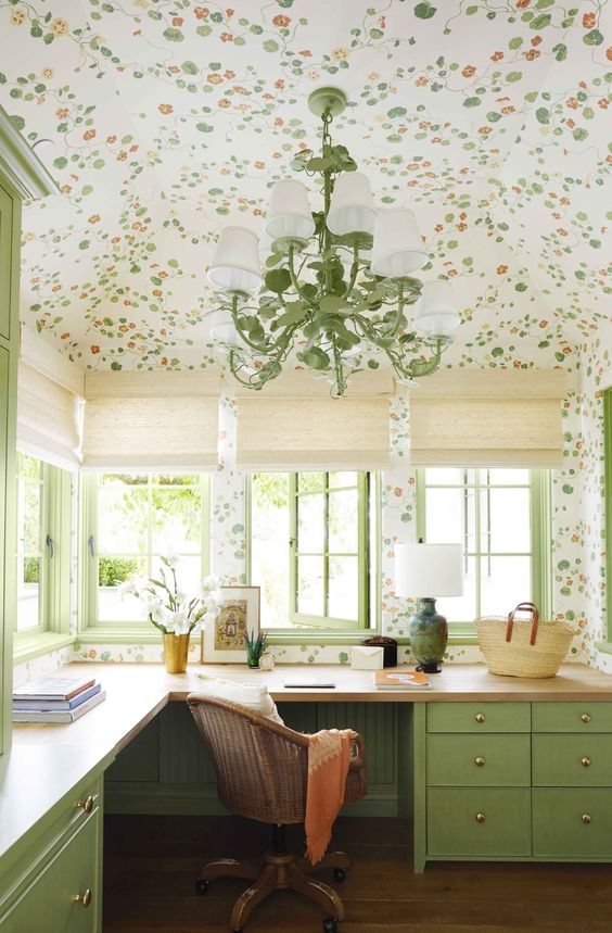 a welcoming home office with a green floral ceiling and walls, a green corner desk, a rattan chair and a green retro chandelier