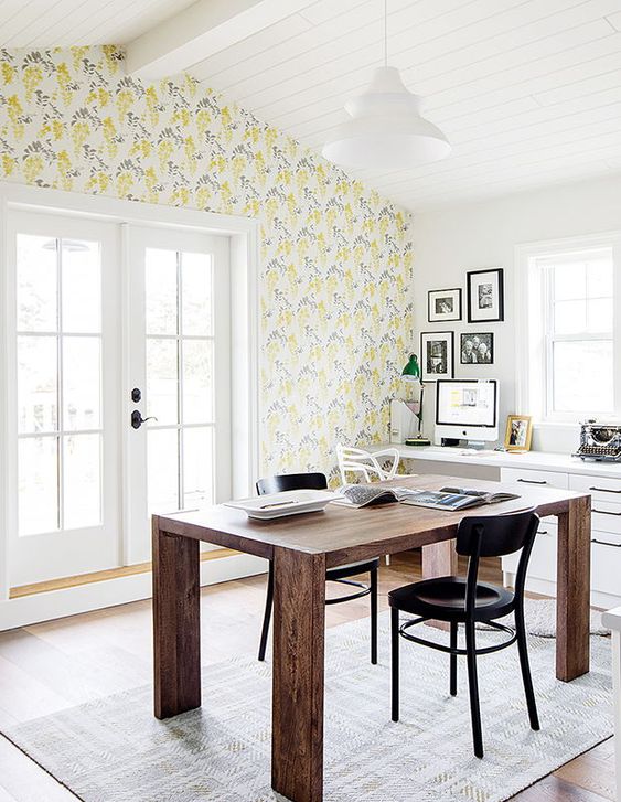 a welcoming spring home office with yellow floral wallpaper, a wooden desk, a gallery wall, black chairs is filled with light