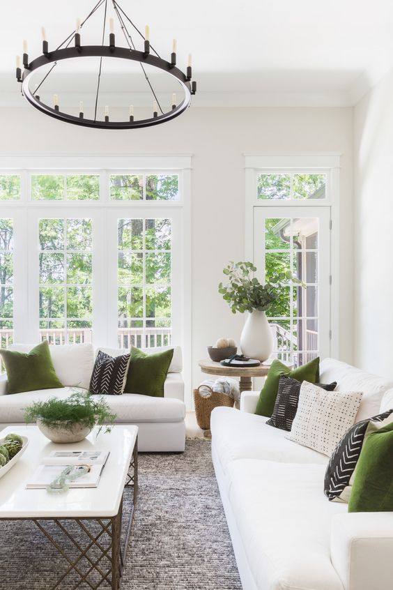 a white living room with elegant and chic furniture, a vintage chandelier, green and printed pillows and greenery in vases