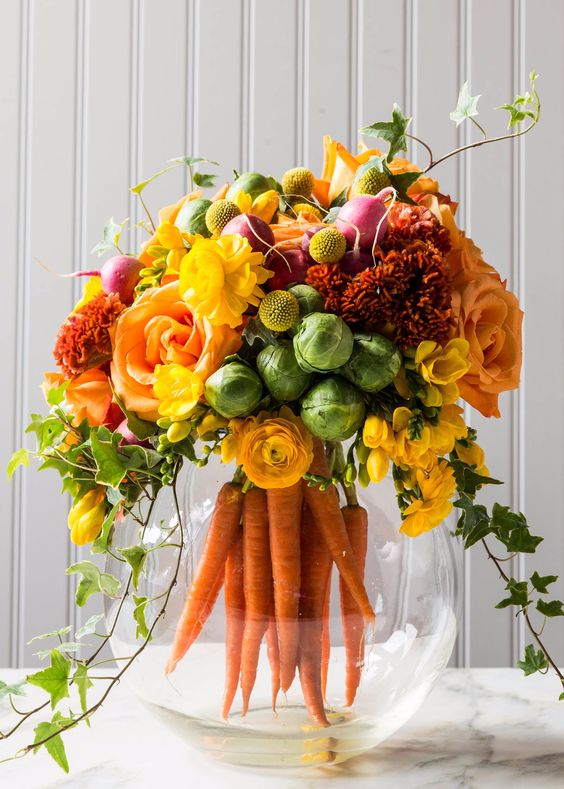 a modern rustic spring centerpiece of an aquarium with carrots, bold blooms and cabbages plus some foliage is a fun idea