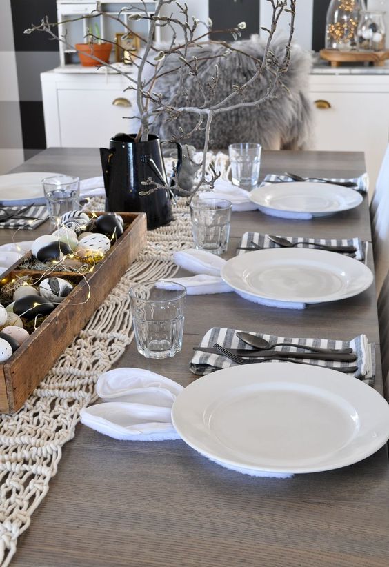a monochromatic Easter tablescape with a macrame runner, a wooden box with dyed eggs and lights, striped napkins and black cutlery