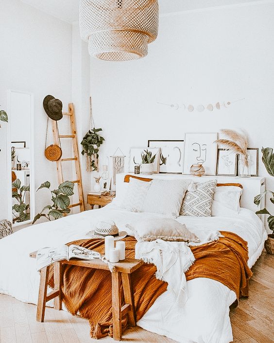 a boho bedroom with a comfy bed, a small wooden bench at the foot, a ladder, a wicker lamp and potted plants