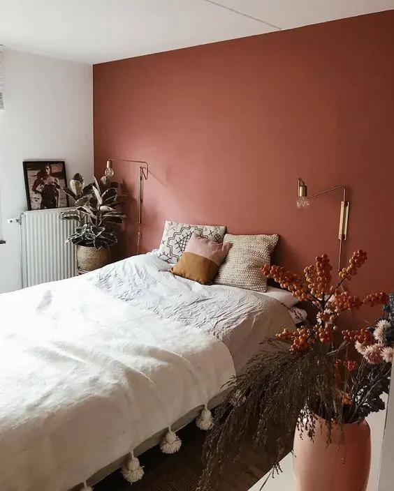 a bold bedroom with a terracotta accent wall, a bed with neutral bedding, gold sconces and potted plants