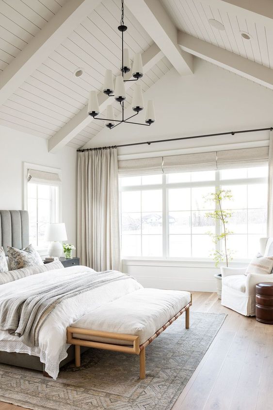 a modern farmhouse neutral bedroom with an upholstered bed, an upholstered bench, a vintage chandelier and a potted plant