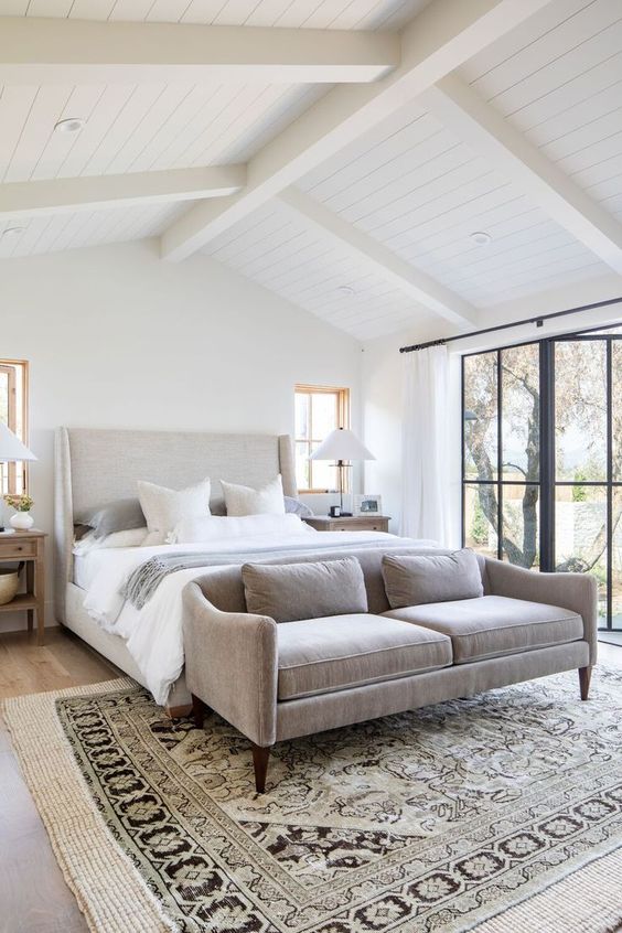 a neutral and welcoming modern farmhouse bedroom with an upholstered bed, a pretty loveseat at the foot, wooden nightstands and lamps