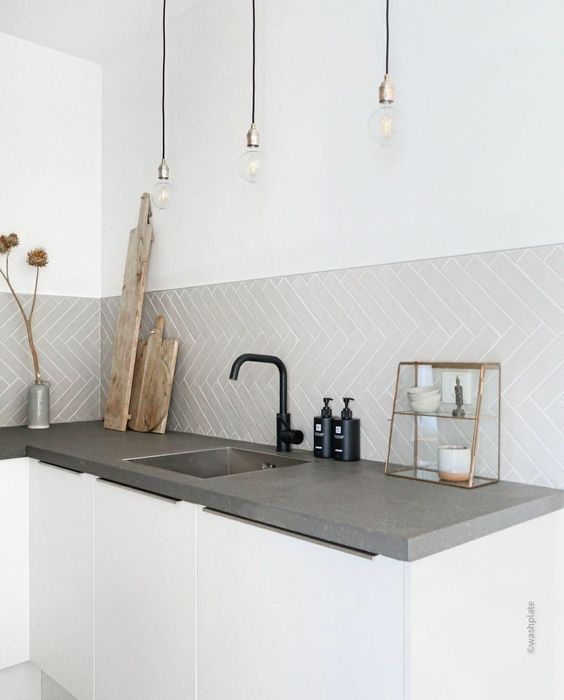 a Scandinavian kitchen with sleke white cabinetry, a concrete countertops, a grey herringbone tile backspplash and pendant bulbs