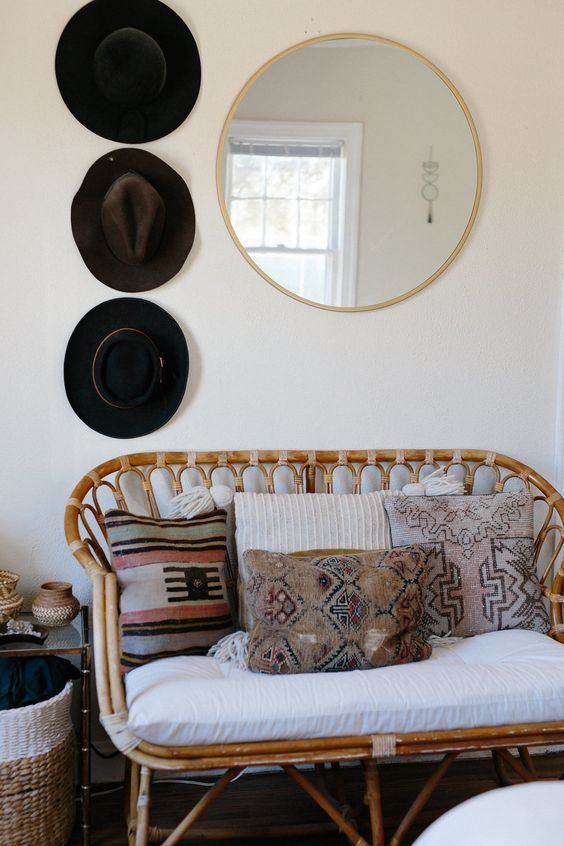 a boho space with a rattan loveseat with boho pillows, a round mirror and hats on display is a chic nook