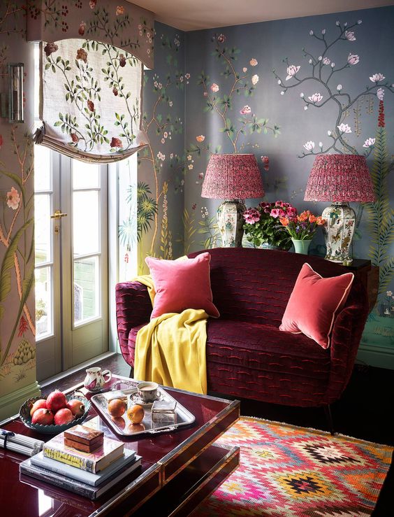 a bright maximalist living room with floral wallpaper, a burgundy loveseat, a colorful ikat rug and floral textiles