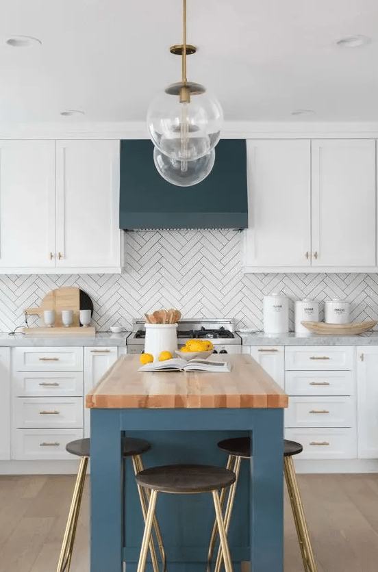 a chic farmhouse kitchen with white shaker cabinets, a herringbone tile backsplash, a blue kitchen island and a hood