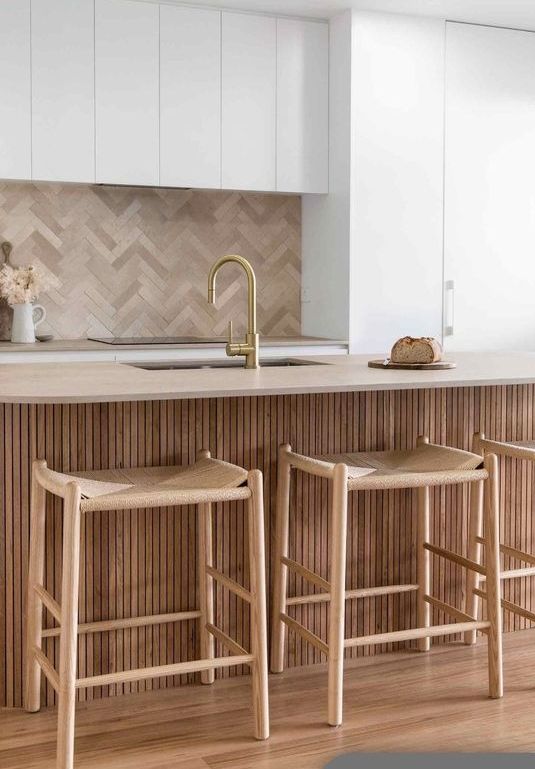 a contemporary kitchen with sleek white cabinets, a kitchen island with fluted texture, woven stools and a beige herringbone tile backsplash
