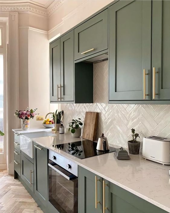 a green farmhouse kitchen with shaker cabinets, white countertops, a white herringbone tile backsplash and gold handles
