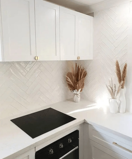 a minimalist white kitchen with shaker cabinets, white herringbone tiles, built in appliances and boho leaves and grasses