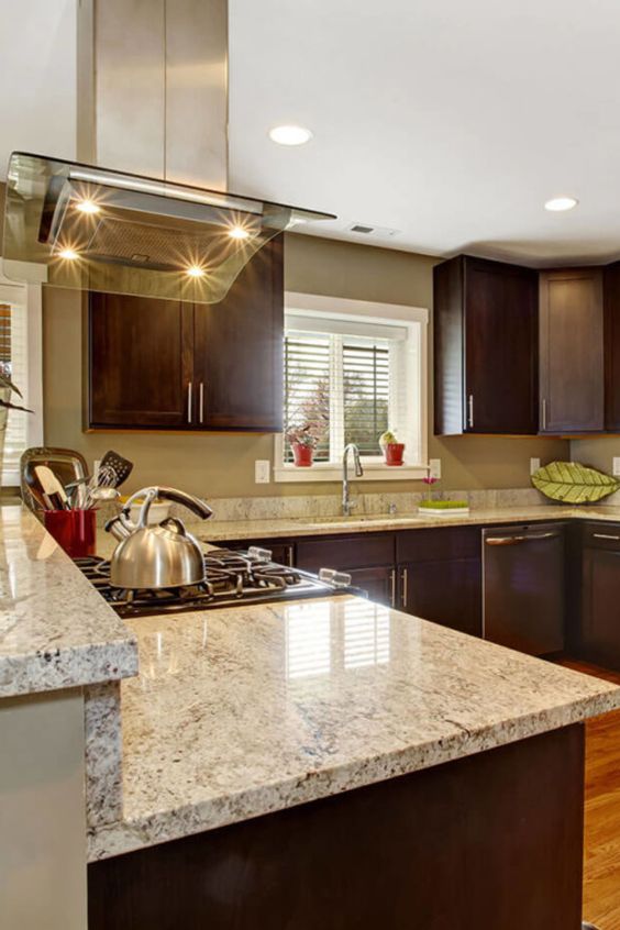 a refined dark kitchen with creamy granite countertops, a stainless steel and glas shood and built-in lights