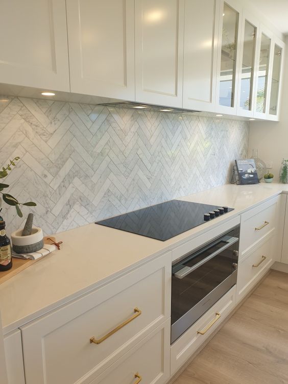 a refined white kitchen with shaker cabinets, a marble herringbone tile backsplash, white stone countertops and built in lights