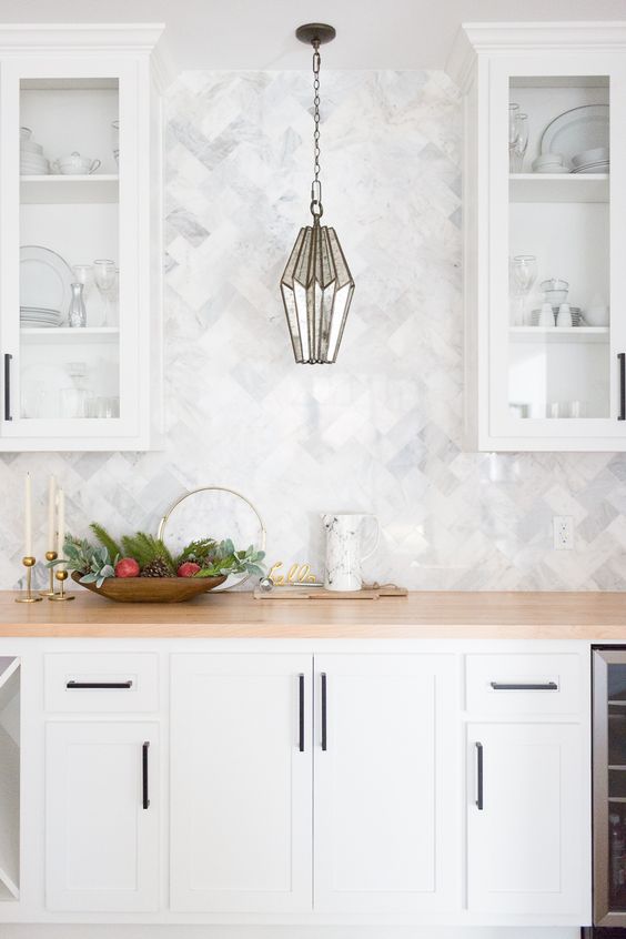 a serene and airy white kitchen with glass upper cabinets and a white herringbone backsplash plus butcherblock countertops