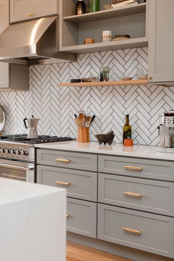 a sophisticated grey farmhouse kitchen with shaker cabinets, a white herringbone tile backsplash, gold fixtures and open shelves