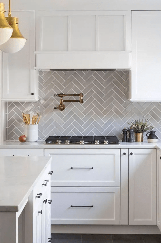 a white farmhouse kitchen with a grey herringbone tile backsplash, a kitchen island, gold pendant lamps