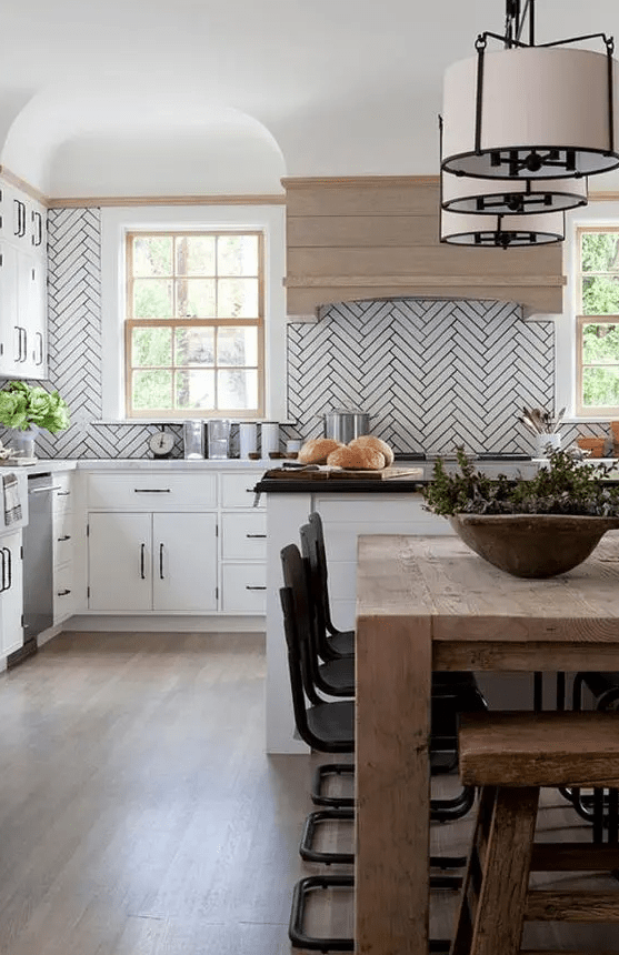 a white farmhouse kitchen with black handles, a white herringbone tile backsplash, a wooden hood, a kitchen island with a black countertop