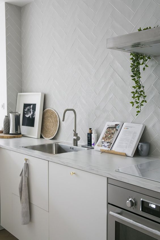 a white kitchen with sleek cabinets, white stone countertops, a white chevron tile wall and some lovely decor