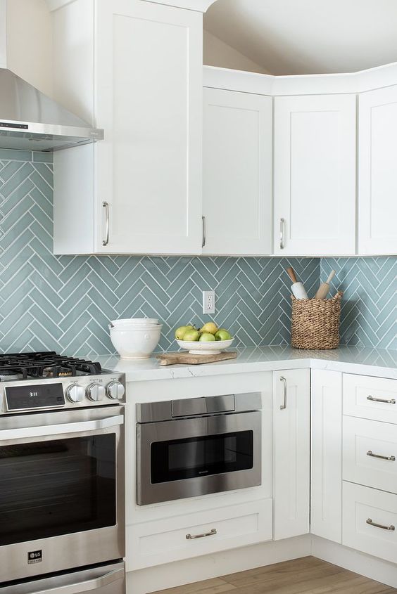 a white shaker style kitchen with white stone countertops and a blue herringbone tile backsplash and stainless steel appliances