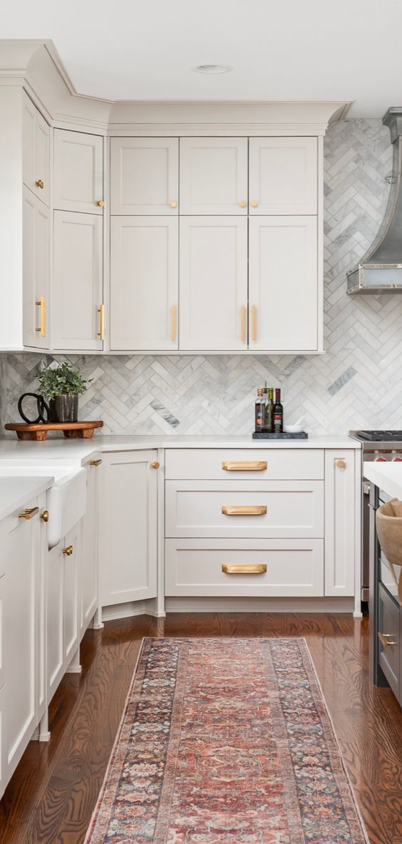 an elegant creamy kitchen with shaker cabinets, a marble herringbone tile backsplash and white stone countertops