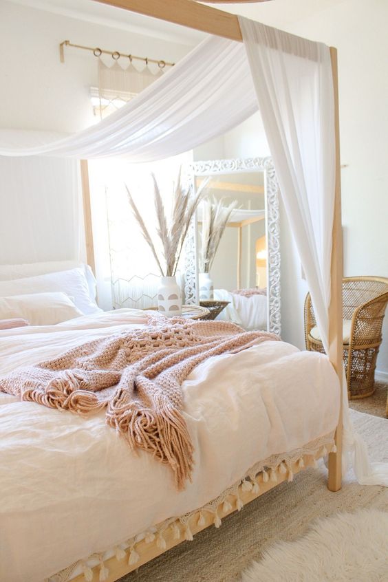 a welcoming and airy bedroom in neutrals and pastels, with a canopy bed with curtains, pastel bedding, a mirror, rattan furniture