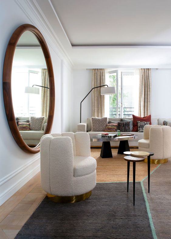 a luxurious neutral living room with an oversized round mirror in a wooden frame, sculptural furniture and bold printed pillows